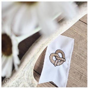 May include: A white fabric banner featuring a detailed embroidered pretzel design in brown and black. The banner is set against a wooden surface, with a blurred white flower and antler in the background.