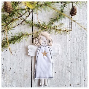 May include: A handmade white felt angel ornament with a gold star on the front. The angel has a smiling face, blonde hair, and white wings. The ornament is hanging on a white wooden surface with pine branches and pine cones.