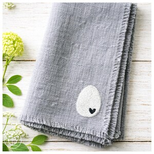 Könnte beinhalten: Eine gefaltete graue Leinen-Serviette mit einem weißen, gestickten Eierdesign und einem kleinen schwarzen Herzen. Die Serviette hat einen Fransenrand und wird auf einer hellen Holzoberfläche präsentiert. Grünes Laub und Blumen sind im Hintergrund.