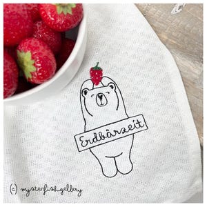 Könnte beinhalten: Weißes Küchentuch mit einem gestickten Bären, der eine Erdbeere auf dem Kopf hält. Der Bär hat das Wort "Erdbärzeit" auf einem Schild. Eine Schüssel mit frischen roten Erdbeeren befindet sich in der oberen linken Ecke.