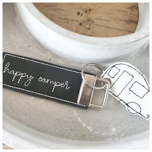May include: Black leather keychain with white stitching that says "happy camper" and a white camper van charm.