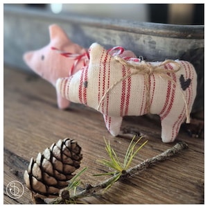 Könnte beinhalten: Zwei handgefertigte Schweineornamente. Eines ist hellrosa, das andere cremefarben mit roten Streifen. Das cremefarbene Schwein hat ein Herzdetail und ist mit Bindfaden gebunden. Ein Tannenzapfen und ein Zweig mit Kiefernnadeln befinden sich im Vordergrund auf einer Holzoberfläche.