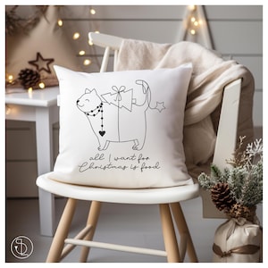 Könnte beinhalten: Ein weißes Kissen mit einer schwarzen Linienzeichnung einer Katze, die einen Weihnachtsbaum auf dem Rücken trägt. Der Text "all I want for Christmas is food" ist unter der Katze aufgedruckt.