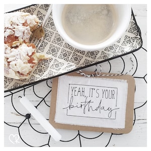 Pode incluir: Um cartão castanho com um inserto de tecido branco. O inserto de tecido tem texto preto que diz "Yeah, it's your birthday". O cartão está sobre uma superfície branca com um design de metal preto.