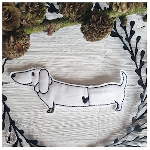 Pode incluir: Um dachshund de feltro branco com detalhes bordados em preto, incluindo um coração no peito. O dachshund está deitado com a cabeça virada para o lado.