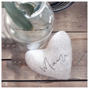 May include: A white felt heart with the word "Mama" written in black cursive script. The heart is sitting on a wooden surface.