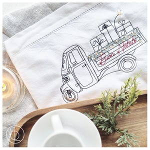 Op de afbeelding: Witte linnen theedoek met een geborduurd ontwerp van een driewielige vrachtwagen met verpakte cadeaus. De vrachtwagen heeft de woorden "Buon Natale" geborduurd op de zijkant. Een takje groen en een theekopje staan op de voorgrond.