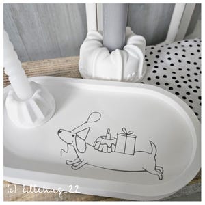 May include: White oval tray featuring a black and white illustration of a dachshund carrying a birthday cake, a present, and a balloon. The tray is on a wooden surface.