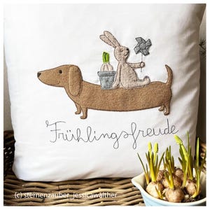 May include: A white pillow with embroidered images of a dachshund, a bunny holding a pinwheel, and a potted plant. The text "Frühlingsfreude" is embroidered below the images.