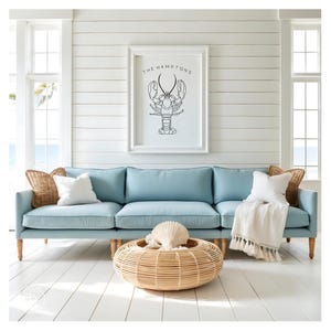 Könnte beinhalten: Ein Wohnzimmer im Küstenstil mit einem hellblauen Sofa, weißen Kissen und einem gewebten Couchtisch. Ein gerahmter Druck mit einer Hummerillustration hängt über dem Sofa. Der Text auf dem Druck lautet "The Hamptons".
