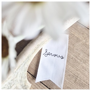May include: A white fabric tag with the word "Serious" embroidered in black cursive script. The tag is placed on a wooden surface, with a blurred white flower and a white antler in the background.