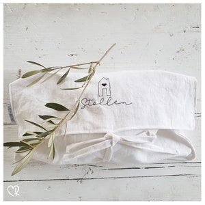 Puede incluir: Bolsa de lino blanca con una casa bordada en negro con un corazón y la palabra "Stollen" debajo. La bolsa está atada con una cinta blanca y una rama de olivo está encima.