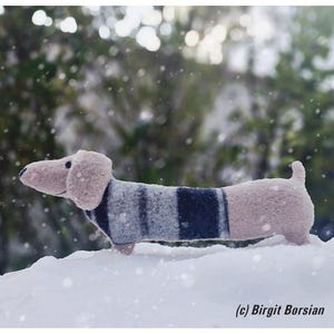 May include: A plush toy dachshund wearing a blue and white striped sweater, standing in the snow. The toy is made of soft, fuzzy fabric and has a pink nose and ears.