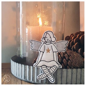 Könnte beinhalten: Ein weißer Filzengel-Anhänger mit schwarzen Stickdetails, darunter Flügel, ein Kleid und Beine. Der Engel sitzt auf einem Metalltablett mit einer Kerze in einem Glasgefäß dahinter.