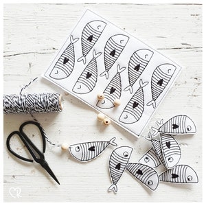 Può includere: Un tessuto bianco con disegni di pesci delineati in nero, ciascuno con un cuore e un motivo a righe. Sono presenti anche un rocchetto di spago bianco e nero, forbici nere e perline di legno, che suggeriscono un progetto fai-da-te.