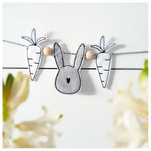 Könnte beinhalten: Eine weiße Schnur-Girlande mit einem grauen Hasen und zwei weißen Karotten. Die Girlande ist mit Holzperlen verziert.