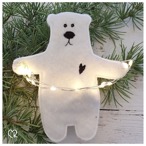 Puede incluir: Adorno de oso polar de fieltro blanco con ojos, nariz y corazón bordados en negro. El oso sostiene una cadena de luces blancas.