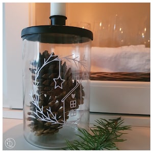 Pode incluir: Um frasco de vidro transparente com tampa preta, cheio de uma pinha e um desenho branco de uma casa com um coração e uma estrela. O frasco está sobre uma superfície branca com um ramo de pinheiro verde em primeiro plano.