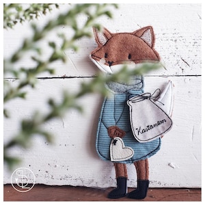 Peut inclure: Un renard en feutre marron portant un manteau en feutre bleu et tenant un sac en feutre blanc avec le mot "Kastanien" brod&eacute; dessus. Le renard est debout sur une surface en bois blanc.