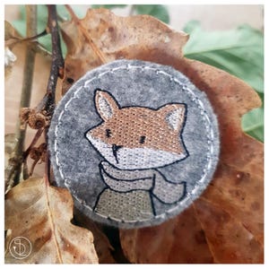 May include: A grey felt circle with a brown fox embroidered on it. The fox is wearing a grey scarf and has a white face.