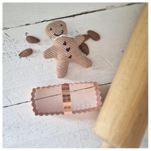 May include: A fabric gingerbread man ornament with brown and white stripes, embroidered facial features, and three small heart buttons. Beside it lies a rectangular copper cookie cutter with scalloped edges and a wooden rolling pin.