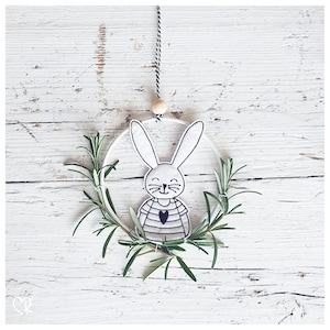 Puede incluir: Un conejito blanco de fieltro con un corazón negro y rayas está bordado en un aro blanco con ramitas de romero verde alrededor.