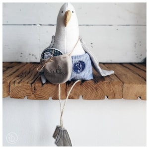 May include: A white stuffed seagull with a blue and white striped fabric on its bottom half. The seagull is holding a small brown bag with the word "beach" embroidered on it. The seagull is sitting on a wooden shelf.