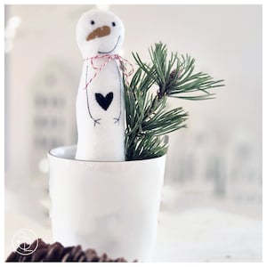 Könnte beinhalten: Eine weiße Schneemannpuppe mit einem aufgenähten schwarzen Herz und Füßen, einer braunen Nase und einer rot-weiß gestreiften Schnur. Der Schneemann befindet sich in einem weißen Becher mit einem Zweig grüner Kiefernnadeln.