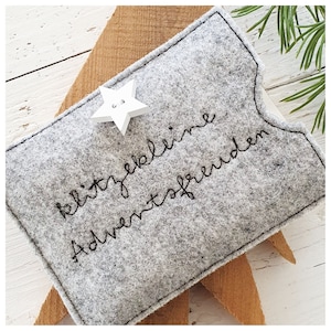 Könnte beinhalten: Grauer Filz-Adventskalender mit einem weißen Stern und dem aufgestickten Text "kleine glitzergeblink Adventssfreuden".