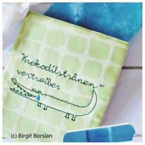 Può includere: Un porta fazzoletti in tessuto verde con un motivo a scacchi. È ricamato con un coccodrillo con il testo "Krokodilstränen-vertreiber" che si traduce in "Scaccia lacrime di coccodrilo".