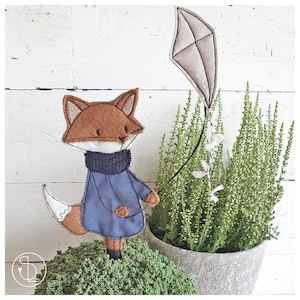 May include: A brown fox wearing a blue jumper and holding a grey kite. The fox is standing on a bed of green moss.