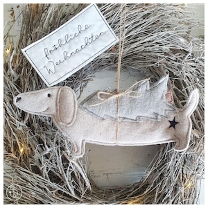 Könnte beinhalten: Ein beigefarbener Filz-Dackel-Anhänger mit einem grauen Filz-Weihnachtsbaum auf dem Rücken. Der Anhänger hängt an einem Zweigkranz mit einem weißen Schild, auf dem "fröhliche Weihnachten" steht.