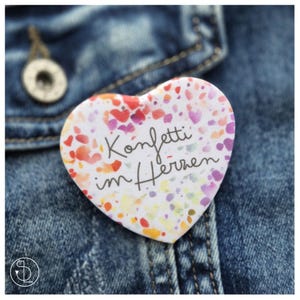 Könnte beinhalten: Ein herzförmiger Button mit buntem Konfetti-Design in Rot, Rosa und Gelb. Der Button trägt den deutschen Text "Konfetti im Herzen" in schwarzer Schrift und ist an einer Jeansjacke befestigt.