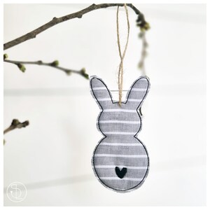 Könnte beinhalten: Ein Stoff-Osterhase mit grau-weißen Streifen und einem schwarzen Herzdetail, der an einer Schnur hängt. Er hängt an einem Ast vor weißem Hintergrund. Der Hase ist aus Stoff gefertigt.