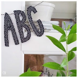 May include: Black and white polka dot fabric letters spelling "ABC" hanging from a string. The letters are hanging in front of a white shelf with a green plant in the background.