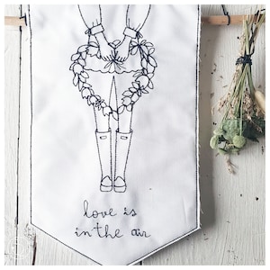 Pode incluir: Uma faixa bordada em preto e branco com uma ilustração simples de uma pessoa segurando uma coroa de flores. A faixa tem um fundo pontiagudo e o texto "love is in the air" está bordado abaixo da ilustração.