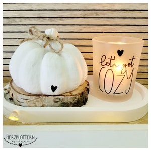 Puede incluir: Calabaza blanca con un lazo de yute sobre una rodaja de madera, junto a un portavelas de vidrio esmerilado con el texto "Let's get cozy" y un corazón negro.