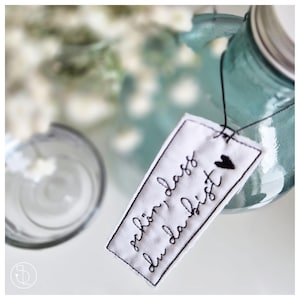 Pode incluir: Uma etiqueta de tecido branco com bordado preto que diz "schön, dass du da bist" e um pequeno coração preto. A etiqueta está presa a um frasco de vidro azul.