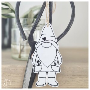 May include: A white gnome-shaped ornament with black embroidered details, including a heart, beard, and pockets with a flower and a tool. The ornament hangs from a natural twine string. A pair of black scissors and a glass jar are in the background.