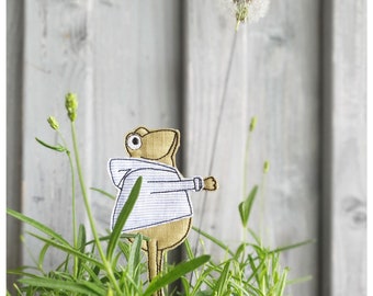 May include: A brown frog wearing a white and blue striped hoodie stands in a pot of green plants. The frog is looking up at a dandelion with a white seed head.