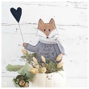 Könnte beinhalten: Ein grauer Filz Fuchs, der einen grauen Schal trägt und einen schwarzen Herzluftballon hält. Der Fuchs ist mit dem Text "Ich liebe Herbst am meisten" bestickt.