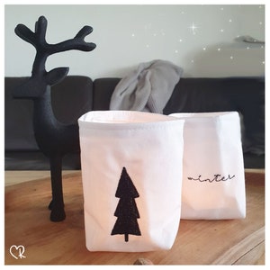 Peut inclure: Deux bougeoirs en tissu blanc avec des broderies noires. Un bougeoir présente un motif d'arbre de Noël stylisé, tandis que l'autre dit "hiver".