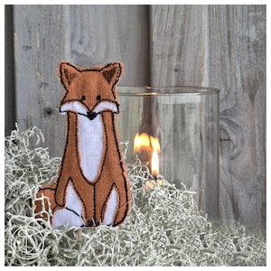Könnte beinhalten: Eine Stoff-Fuchsfigur in Braun und Weiß mit schwarzen Nähten. Der Fuchs sitzt auf getrocknetem Moos, mit einer Kerze in einem Glasbehälter im Hintergrund. Der Hintergrund ist eine Holzwand.