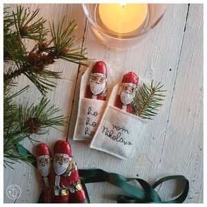 Puede incluir: Dos pequeñas bolsas de fieltro blanco con figuras de chocolate de Papá Noel en su interior. Las bolsas están bordadas con "ho ho ho" y "vom Nikolaus".