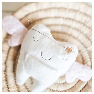 Può includere: Un dente di peluche bianco con un volto sorridente, dettagli cuciti in nero e una stella dorata. Il dente ha due piccole ali rosa con un motivo a pois. Lo sfondo è un materiale beige tessuto.