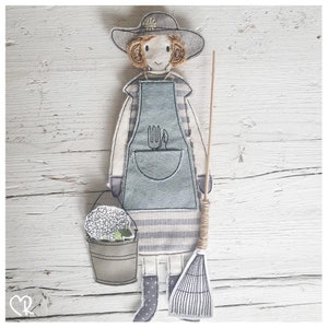 May include: A handmade fabric doll wearing a blue and white striped dress and a blue apron with a gardening tool design. The doll is holding a bucket with white flowers and a rake.