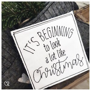 May include: White fabric square with black embroidered text that reads "It's beginning to look a lot like Christmas".