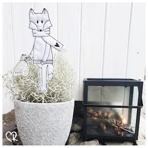 May include: A white felt fox with black embroidered details, wearing a scarf with a heart, holding a small white bag with the word "Knus" on it. The fox is standing in a white pot filled with white dried moss. A black metal lantern with a candle inside is in the background.