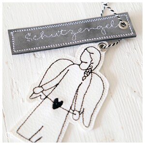May include: A white felt angel ornament with black embroidery, holding a small black heart. The angel is attached to a grey leather tag with the word "Schutzengel" embroidered in white.