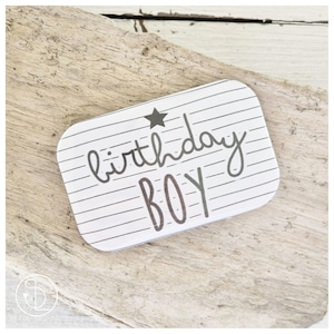 Könnte beinhalten: Ein rechteckiges Etikett mit weiß-grauen Streifen und dem Schriftzug "birthday BOY" in handschriftlicher Schrift. Über dem Wort "birthday" befindet sich ein kleiner schwarzer Stern. Das Etikett liegt auf Treibholz.